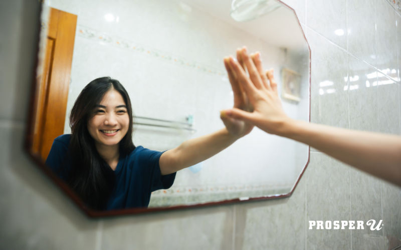 Young Asian woman in a blue t-shirt smiles at herself in the mirror with her left-arm extended out so her hand is touching the mirrored version of her hand. She just finished saying mindful morning mantras.